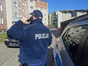 Policjant stojący tyłem przy samochodzie z ręką uniesioną z telefonem przy uchu, przed blokiem
