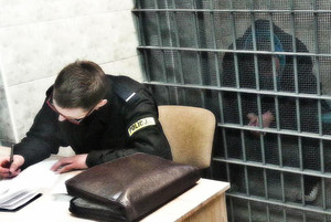 Zatrzymana osoba w celi i policjant siedzący na krześle za biurkiem przed celą